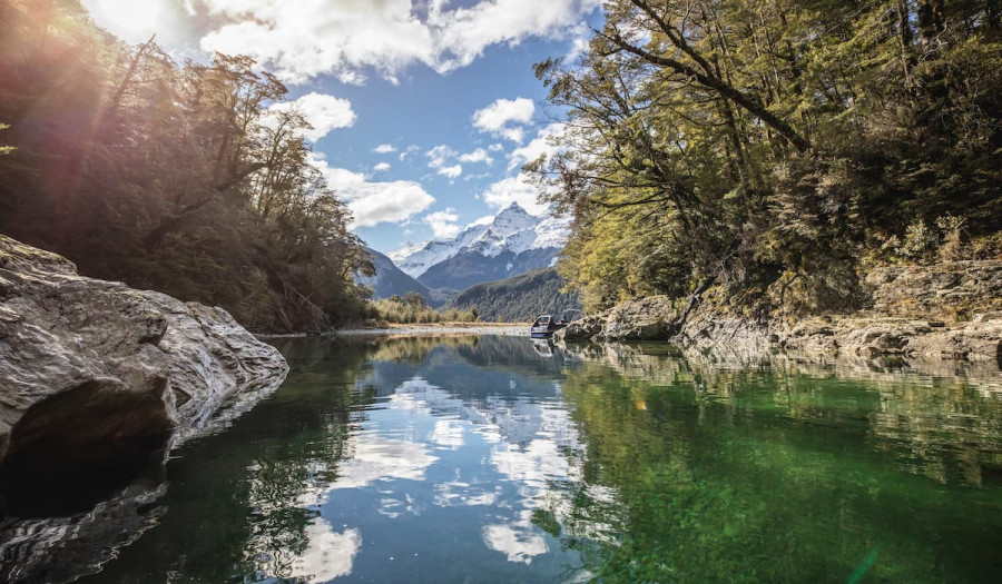 Still water on the Dart River reflecting surrounding trees, with mountains in the distance